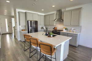 Kitchen with gray cabinetry, a breakfast bar, wall chimney range hood, a kitchen island with sink, and range with gas stovetop