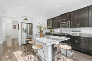 Kitchen featuring light stone counters, stainless steel appliances, a kitchen island with sink, light wood finished floors, and recessed lighting