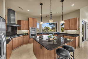 Kitchen featuring wood finish cabinets, dark stone counters, decorative light fixtures, a kitchen island with sink, and stainless steel appliances