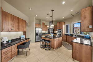 Kitchen with a breakfast bar area, decorative light fixtures, dark stone counters, brown cabinetry, and appliances with stainless steel finishes