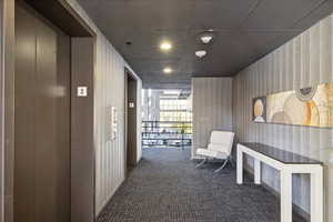 Hallway featuring elevator, wallpapered walls, dark colored carpet, expansive windows, and recessed lighting