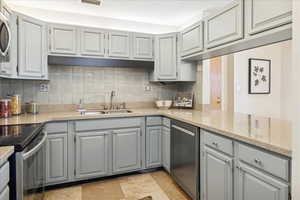 Kitchen featuring gray cabinetry, appliances with stainless steel finishes, tile backsplash, and light stone countertops. Doorway to hallway leading to emergency exit visible.