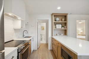 Kitchen with appliances with stainless steel finishes, tasteful backsplash, light wood-type flooring, open shelves, and wall chimney range hood