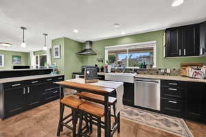 Kitchen with dark cabinets, stainless steel appliances, wall chimney range hood, light stone counters, and recessed lighting