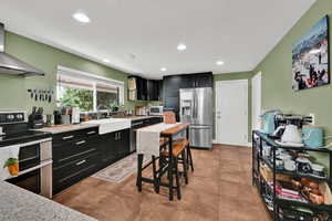 Kitchen with dark cabinetry, stainless steel appliances, range hood, light tile patterned floors, and recessed lighting
