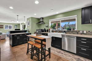 Kitchen with dark cabinetry, decorative light fixtures, appliances with stainless steel finishes, wall chimney range hood, and light stone counters