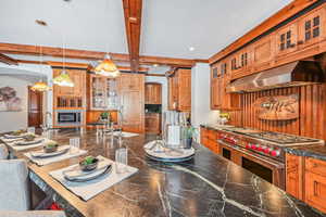 Kitchen with glass insert cabinets, stainless steel appliances, range hood, dark countertops, and pendant lighting