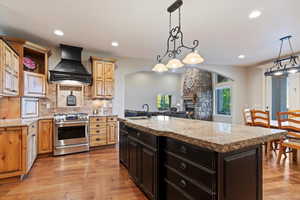 Kitchen with stainless steel gas range, a center island with sink, custom exhaust hood, backsplash, and a stone fireplace