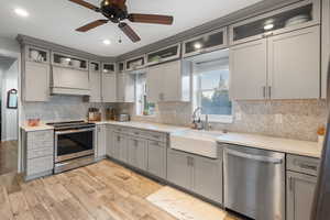 Kitchen featuring gray cabinetry, stainless steel appliances, custom exhaust hood, light wood-style flooring, and tasteful backsplash