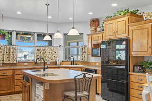 Kitchen featuring black appliances, light tile patterned floors, a kitchen island with sink, hanging light fixtures, and tile counters
