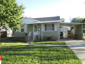 Bungalow-style home with a front lawn, concrete driveway, an attached carport, roof with shingles, and an outbuilding