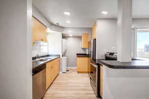 Kitchen with light brown cabinets, stainless steel appliances, light wood-style floors, dark countertops, and washer / clothes dryer