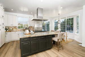 Kitchen with tasteful backsplash, light stone counters, light wood-type flooring, island range hood, and dark cabinetry