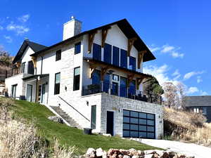 View of side of property featuring a balcony, stone siding, a garage, driveway, and a chimney