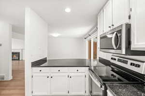 Kitchen featuring stainless steel appliances, white cabinets, light wood finished floors, dark stone countertops, and a glass covered fireplace