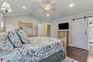 Primary bedroom featuring ornamental molding, vaulted ceiling, a barn door, wood finished floors, and a ceiling fan