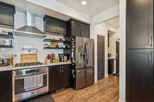 Kitchen with open shelves, stainless steel appliances, wall chimney exhaust hood, light wood-style flooring, and decorative backsplash
