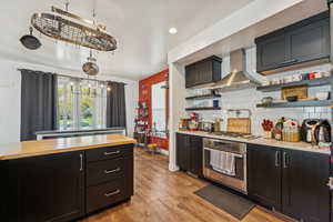 Kitchen with dark cabinets, wall chimney exhaust hood, appliances with stainless steel finishes, open shelves, and light wood-type flooring