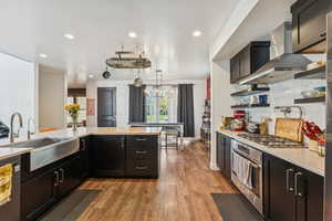 Kitchen with dark cabinetry, light wood finished floors, appliances with stainless steel finishes, wall chimney range hood, and recessed lighting