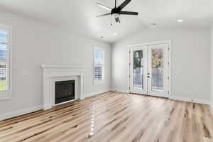 Unfurnished living room featuring a brick fireplace, light wood finished floors, lofted ceiling, ceiling fan, and french doors