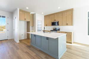 Kitchen featuring decorative backsplash, appliances with stainless steel finishes, an island with sink, light wood-style flooring, and light stone counters