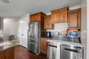 Kitchen with stainless steel appliances, brown cabinets, dark wood-style floors, dark stone counters, and a textured ceiling
