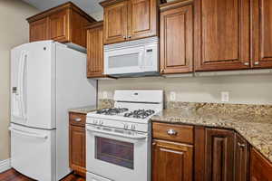 Kitchen featuring white appliances, brown cabinetry, light stone countertops, and dark wood-type flooring