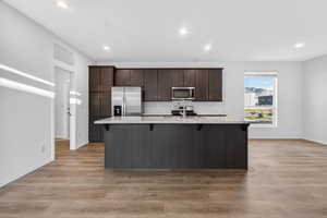 Kitchen featuring dark brown cabinets, a kitchen bar, light stone countertops, an island with sink, and appliances with stainless steel finishes