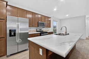 Kitchen featuring stainless steel appliances, a kitchen island with sink, light wood-type flooring, brown cabinetry, and recessed lighting