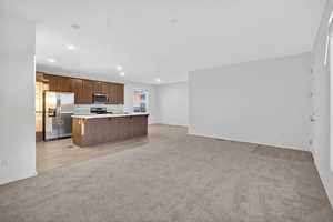 Kitchen featuring open floor plan, light carpet, light countertops, appliances with stainless steel finishes, and a kitchen island with sink