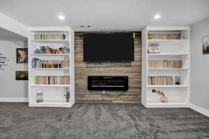 Living area featuring built in shelves, wood-clad back wall, dark carpet, a textured ceiling, and an electric fireplace