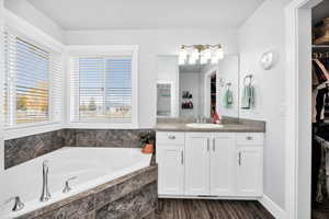 Primary suite bathroom featuring a spacious closet, vanity, an oversized corner tub with tile surround, and dark wood-style flooring