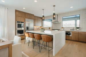 Kitchen with light wood-style floors, pendant lighting, stainless steel appliances, light stone countertops, and wall chimney range hood