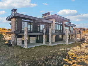 Rear view of house with stone siding, a chimney, a balcony, and a patio