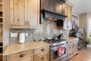 Kitchen with gas range, decorative backsplash, premium range hood, light stone countertops, and light wood finished floors