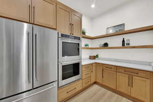 Kitchen with open shelves, stainless steel appliances, light brown cabinets, light wood finished floors, and light stone countertops