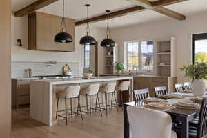 Open-concept kitchen and dining area featuring a large waterfall island with barstool seating, natural wood-finish flooring, exposed ceiling beams, a pot filler faucet, and built-in shelving.