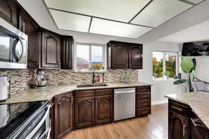 Kitchen with dark brown cabinets, light wood-type flooring, stainless steel appliances, light stone countertops, and backsplash