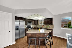 Kitchen featuring dark brown cabinetry, appliances with stainless steel finishes, a kitchen island, a breakfast bar area, and light stone counters