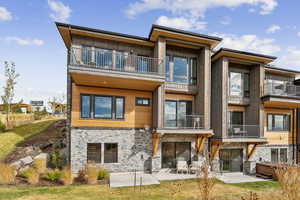 Rear view of property featuring a balcony, a hot tub, a patio, stone siding, and a lawn