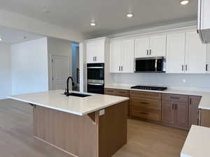 Kitchen with light wood-style flooring, a kitchen island with sink, brown cabinetry, recessed lighting, and white cabinets