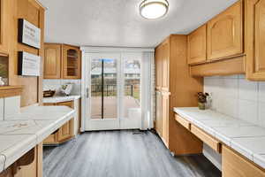 Kitchen featuring tasteful backsplash, a textured ceiling, wood finished floors, tile countertops, and glass insert cabinets