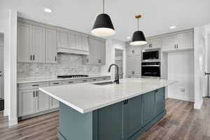 Kitchen with decorative backsplash, stainless steel appliances, light wood-type flooring, light stone countertops, and an island with sink