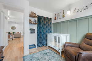 Bedroom with a nursery area, a decorative wall, wood finished floors, and a wainscoted wall