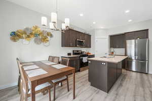 Kitchen with dark brown cabinets, stainless steel appliances, an island with sink, decorative light fixtures, and light wood-style flooring