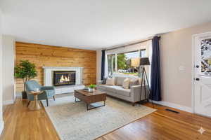 Digitally staged. Living room featuring wooden walls, a brick fireplace, and light wood-style floors