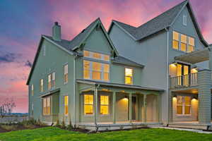 Back of property at dusk featuring roof with shingles, a chimney, stucco siding, a balcony, and a yard