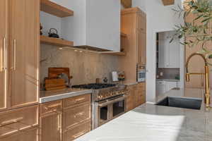 Kitchen with decorative backsplash, appliances with stainless steel finishes, wall chimney exhaust hood, open shelves, and brown cabinetry
