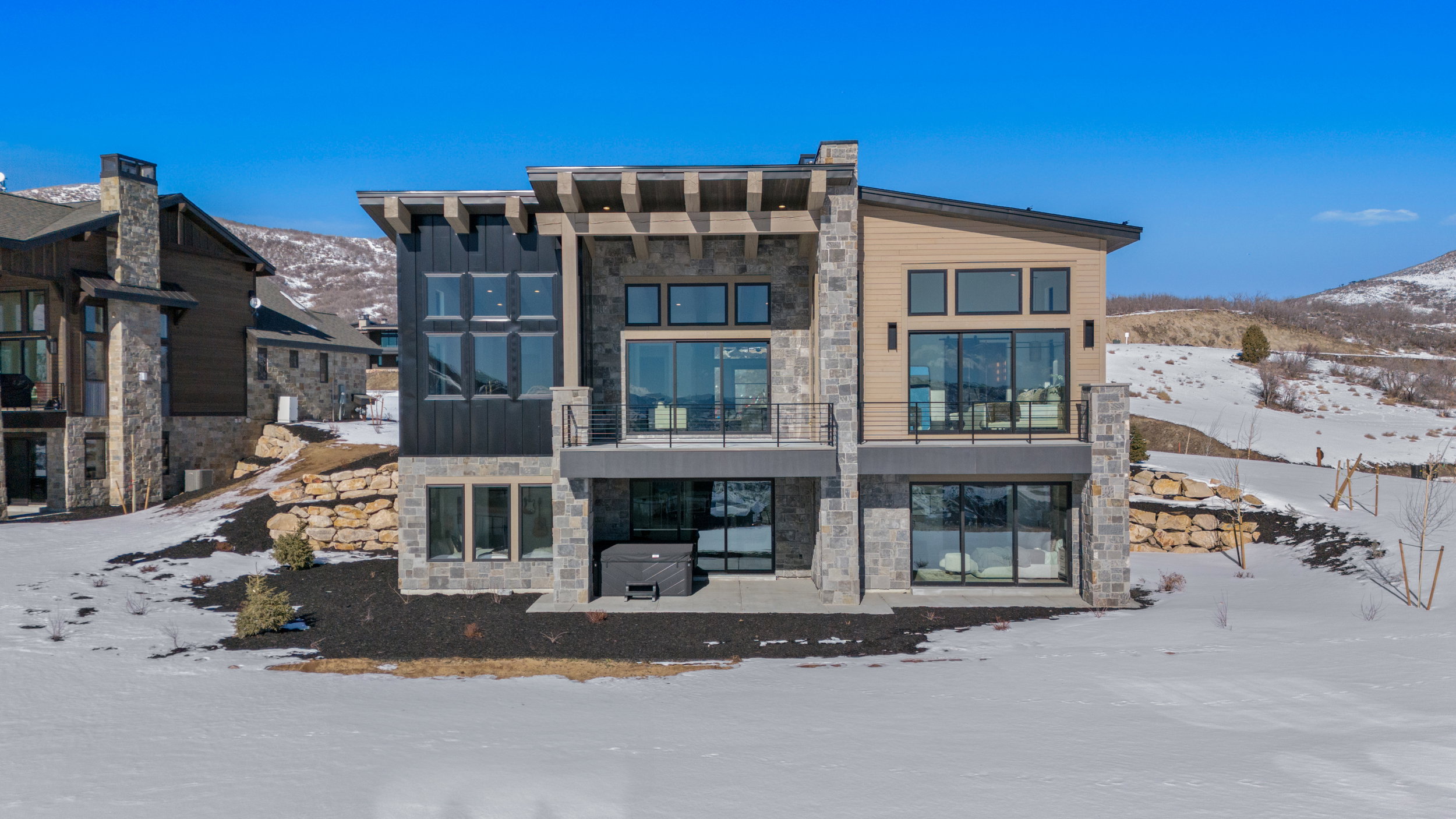 Snow covered back of property featuring stone siding, a patio, a mountain view, and a balcony