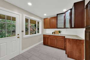 Kitchen with light stone counters, decorative backsplash, brown cabinetry, recessed lighting, and glass insert cabinets